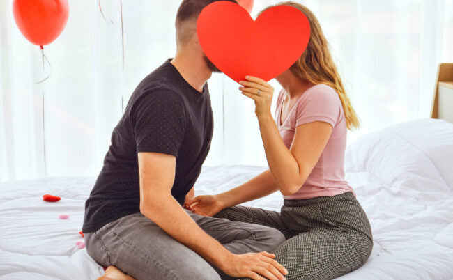 Un homme et une femme assis face à face qui discutent le lubrifiant intime ensemble, le visage caché derrière un grand cœur rouge