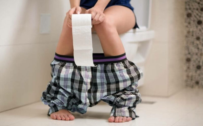 Dranginkontinenz - Frau auf Toilette mit Toilettenpapier und heruntergelassener Hose - Frau mit Blasenschwäche - DE